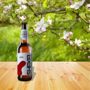Botella de cerveza Shekel de Cervecería Tenek, estilo ámbar con pirul, sobre una mesa de madera con flores de fondo.