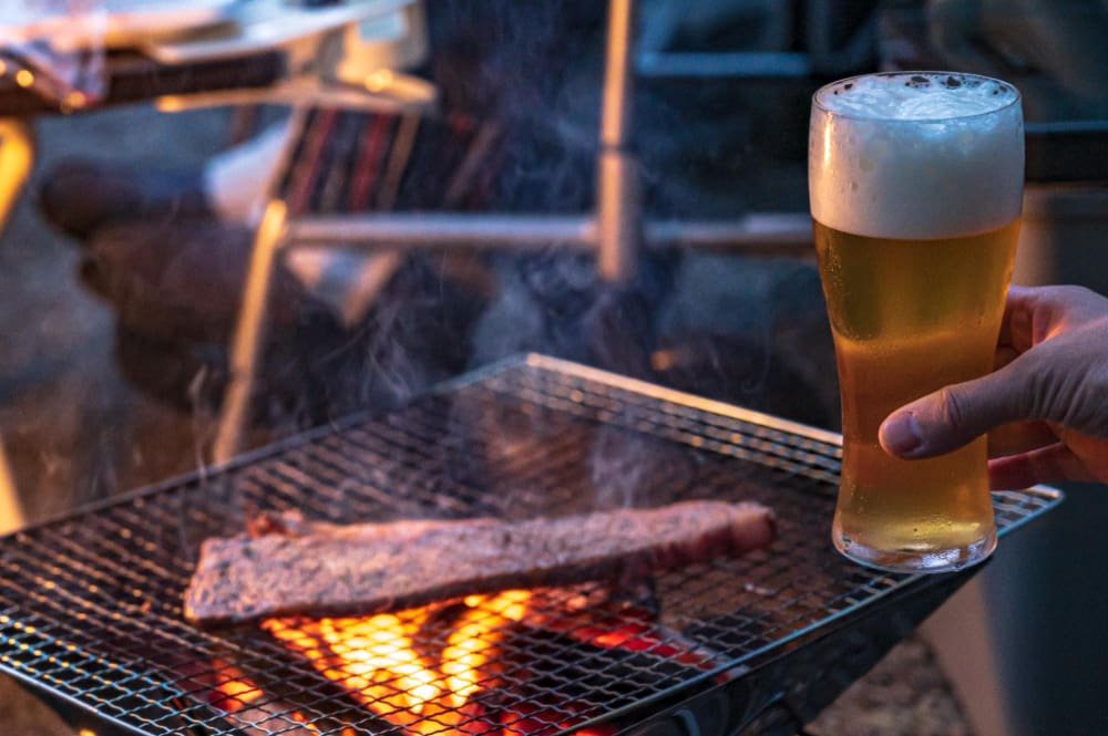 Vaso especial para cerveza artesanal lleno, con una capa de espuma blanca en la parte superior, en la mano de un hombre que está junto a una parrilla con carne asada en Monterrey