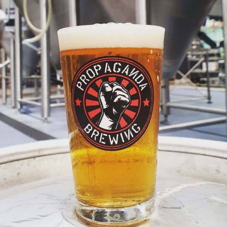 Vaso de cerveza rubia de Propaganda Brewing con la cervecería de fondo.